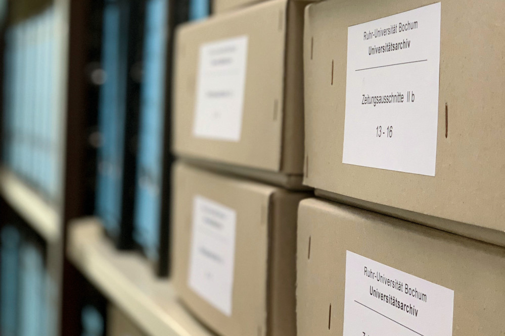 Die Ansicht zeigt ein Archivregal mit etikettierten Aktenkartons und Aktenordnern in einem der Magazinräume
