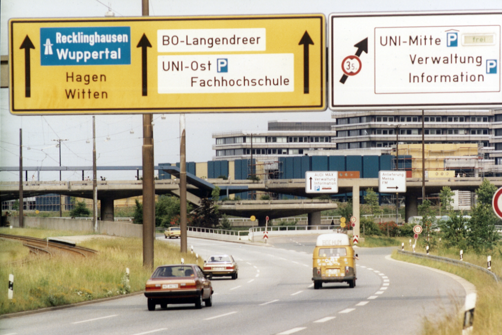 Die Farbfotografie zeigt eine Ansicht der Universitätsstraße in Richtung Langendreer/Witten in den 1970er Jahren. Links neben der Straße sind die Straßenbahngleise zu sehen. Im Hintergrund von links nach rechts befinden sich die Unibrücke, die im Bau befindlichen Werkhallen vor der I-Gebäudereihe.
