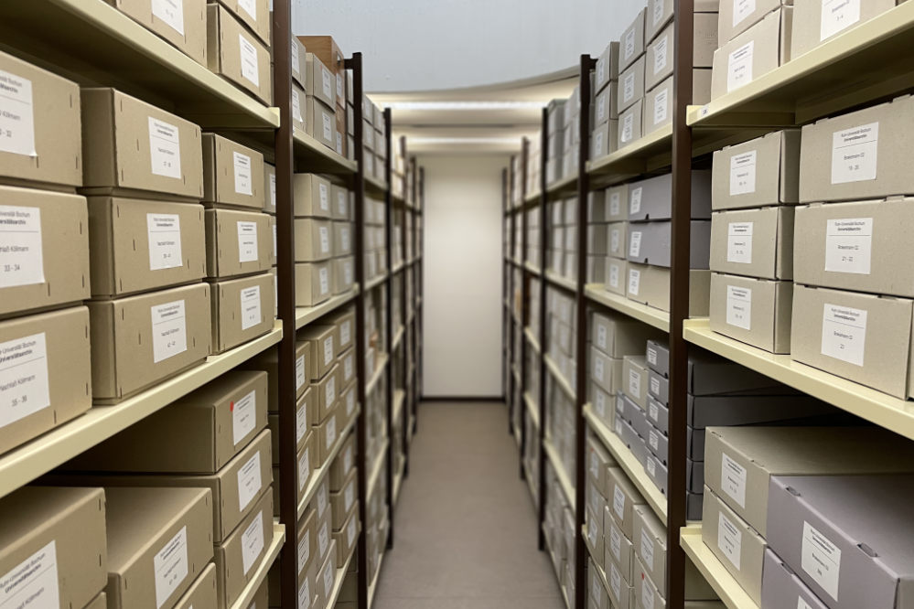 Blick in den Gang eines Magazinraums des Universitätsarchivs. Links und rechts sind volle Regale mit Aktenordnern und Archivkisten zu sehen.
