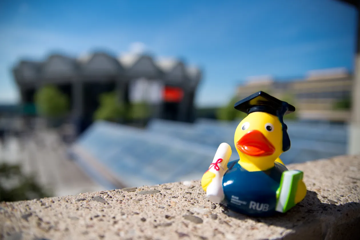 Ein Foto von einer Quitscheente mit Doktorhut auf einer Mauer. Im Hintergrund ist verschwommen das Audimax und das Gebäude GA zu sehen.