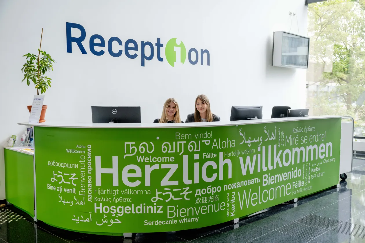 Foto: Hinter einer grünen Theke mit dem weißen Schritfzug "Herzlich willkommen" in vielen unterschiedlichen Sprachen und Schriften sind zwei sitzende Personen und drei Monitore zu sehen. Auf dem linken Ende der Theke steht eine Pflanze. An der Wand steht der Schriftzug "Reception" und es hängt ein Monitor an der Wand.