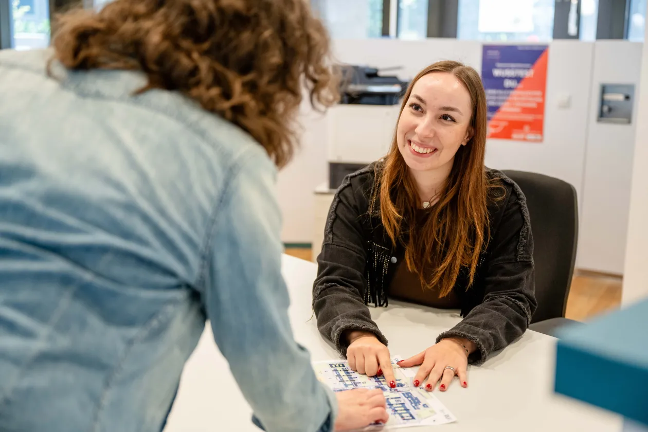 Foto: Eine Person sitzt hinter einer Theke, man sieht sie von vorne. Eine andere Person steht vor der Theke, man sieht ihren Rücken. Beide Personen zeigen auf eine Karte des RUB-Campus, die auf der Theke liegt. Die Person hinter der Theke schaut die andere Person lächelnd an.