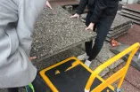Zwei stark abgeschnittene Personen heben eine Waschbetonplatte, wie sie für die RUB typisch sind, an. Unten rechts im Bild steht bereits ein gelber Handkarren bereit um die Platte abzutransportieren.