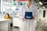 Foto: Frau im weißen Kittel steht in einem Labor und hält lächelnd einen RUB-Kubus vor sich. 