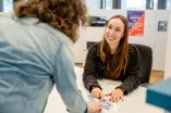 Foto: Eine Person sitzt hinter einer Theke, man sieht sie von vorne. Eine andere Person steht vor der Theke, man sieht ihren Rücken. Beide Personen zeigen auf eine Karte des RUB-Campus, die auf der Theke liegt. Die Person hinter der Theke schaut die andere Person lächelnd an.