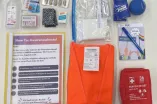 Verschiedene Materialen liegen auf einem weißen Hintergrund. Zu sehen sind eine Mappe, eine Warnweste, eine Erste-Hilfe-Tasche, Hygiene- und Schreibutensilien.