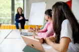 Foto: Seminar in einem Seminarraum. Zwei Personen sitzen und schauen zur Seminarleitung, die an einer Flipchart steht.