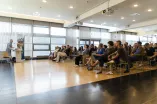 Foto: Vortrag im Veranstaltungszentrum. Eine Person steht hinter dem Rednerpult, die anderen sitzen und gucken die Person an.