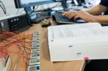 Foto: Auf einem Schreibtisch befindet sich ein aufgeschlagenes Buch, Platinen und Kabelgewirr sowie im Hintergrund auf einer PC-Tastatur tippende Hände.pende 
