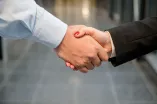 Das Bild zeigt einen Handschlag zwischen einer kräftigeren Hand mit Hemdsärmel und einer Hand mit einem schwarzen Blazer-Ärmel und rot lackierten Nägeln