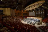 Photo: View from the upper tiers of the Audimax toward the stage, where the choir and orchestra are performing at the 2026 semester concert. The stage is lit. The well-filled seating sections are darkened.