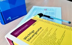 Bücher Steuergesetze und Hochschulrechnungslegung liegen neben Formular und Kulli auf einem Tisch. 