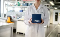 Foto: Frau im weißen Kittel steht in einem Labor und hält lächelnd einen RUB-Kubus vor sich. 