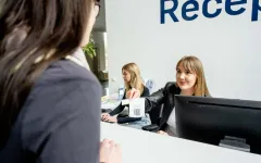 Foto: Zwei Personen sitzen hinter einer Theke, eine Person steht vor der Theke. Eine Person hinter der Theke schreibt auf einer Tastatur, die andere Person hinter der Theke überreicht lächeldn eine Wartemarke.