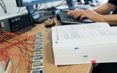 Foto: Auf einem Schreibtisch befindet sich ein aufgeschlagenes Buch, Platinen und Kabelgewirr sowie im Hintergrund auf einer PC-Tastatur tippende Hände.pende 
