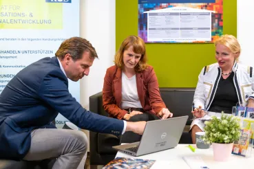 Drei Personen in einer Sitzgruppe schauen und zeigen auf den Laptop-Bildschirm.