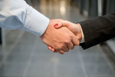 Das Bild zeigt einen Handschlag zwischen einer kräftigeren Hand mit Hemdsärmel und einer Hand mit einem schwarzen Blazer-Ärmel und rot lackierten Nägeln