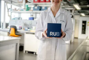 Foto: Frau im weißen Kittel steht in einem Labor und hält lächelnd einen RUB-Kubus vor sich. 