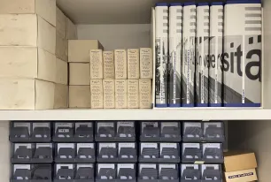 Auf zwei Regalbrettern befinden sich diverse Diasammlungen in transparenten Boxen, eine Publikationsreihe, deren Buchrücken zusammen ein Foto, auf dem der Schriftzug "Universität" zu sehen ist, ergeben, sowie diverse kleinere Materialien in Kartonagen zu sehen.