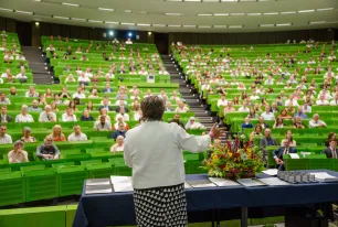 Foto: Gut gefüllter Hörsaal mit grünen Sitzbänken. Vorne steht die Kanzlerin, neben ihr ein großer Blumenstrauß.