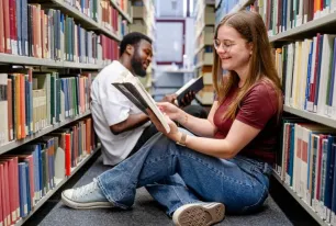 Zwei Studierende sitzen in der Bibliothek auf dem Boden und lesen