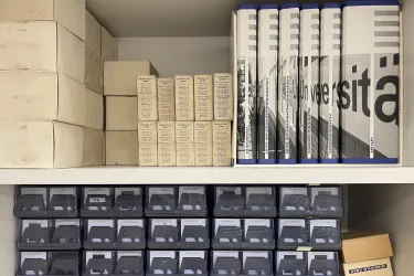 Auf zwei Regalbrettern befinden sich diverse Diasammlungen in transparenten Boxen, eine Publikationsreihe, deren Buchrücken zusammen ein Foto, auf dem der Schriftzug "Universität" zu sehen ist, ergeben, sowie diverse kleinere Materialien in Kartonagen zu sehen.