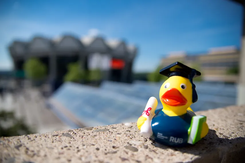 Ein Foto von einer Quitscheente mit Doktorhut auf einer Mauer. Im Hintergrund ist verschwommen das Audimax und das Gebäude GA zu sehen.