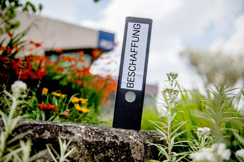Ein mit dem Wort Beschaffung beschrifteter Aktenordnerrücken steht im Freien senkrecht auf einem Stein und ist umgeben von Blumen, im Hintergrund der RUB-Würfel.