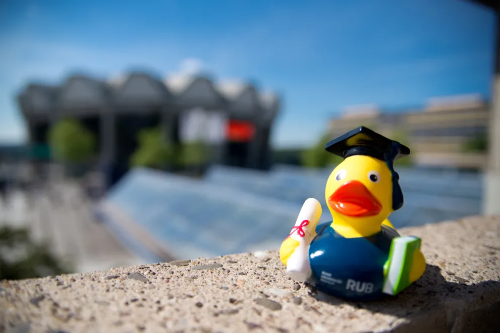 Ein Foto von einer Quitscheente mit Doktorhut auf einer Mauer. Im Hintergrund ist verschwommen das Audimax und das Gebäude GA zu sehen.