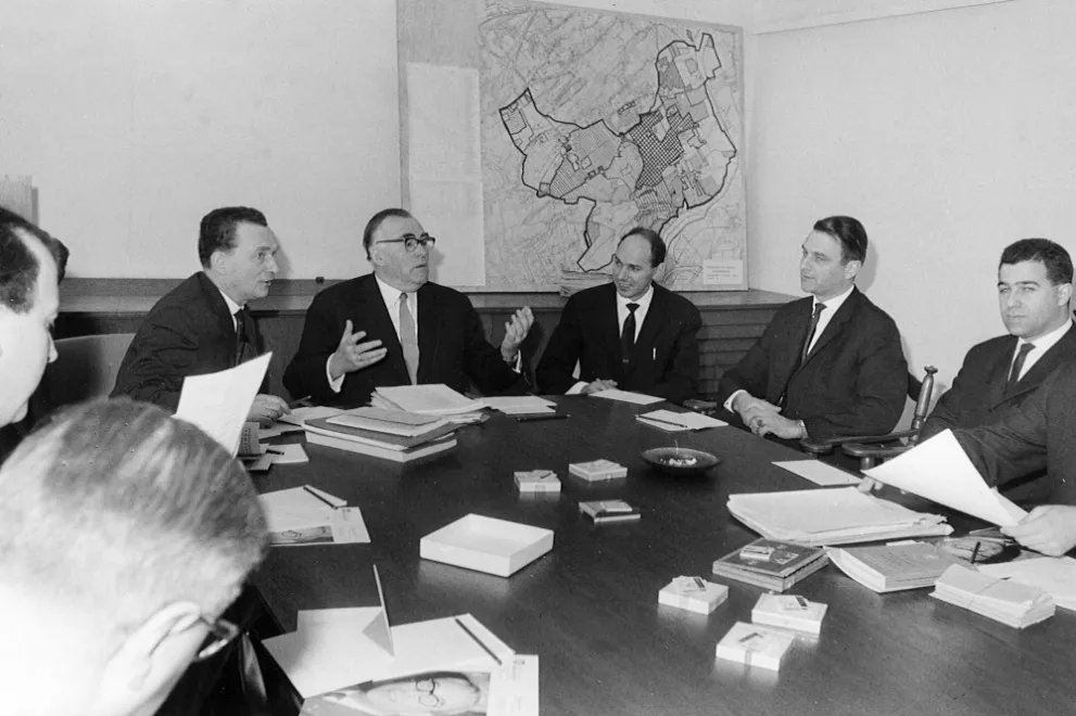 Schwarzweiß-Fotografie der zweiten Sitzung der Gesamtheit der Professoren der RUB am 24.2.1964 im Sitzungssaal der Universitätsverwaltung; am Kopfende des Tisches Prof. Wenke (gestikulierend), links davon Kanzler Seel. Im Hintergrund an der Wand ist als großformatiger Ausdruck "Die Henne" zu sehen: Eine Karte des Suchgebiets für den Standort der RUB im Bochumer Süden, dessen äußere Kontur an eine abstrakte Henne erinnert.