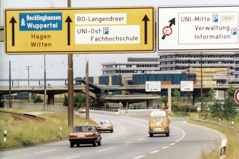 Die Farbfotografie zeigt eine Ansicht der Universitätsstraße in Richtung Langendreer/Witten in den 1970er Jahren. Links neben der Straße sind die Straßenbahngleise zu sehen. Im Hintergrund von links nach rechts befinden sich die Unibrücke, die im Bau befindlichen Werkhallen vor der I-Gebäudereihe.