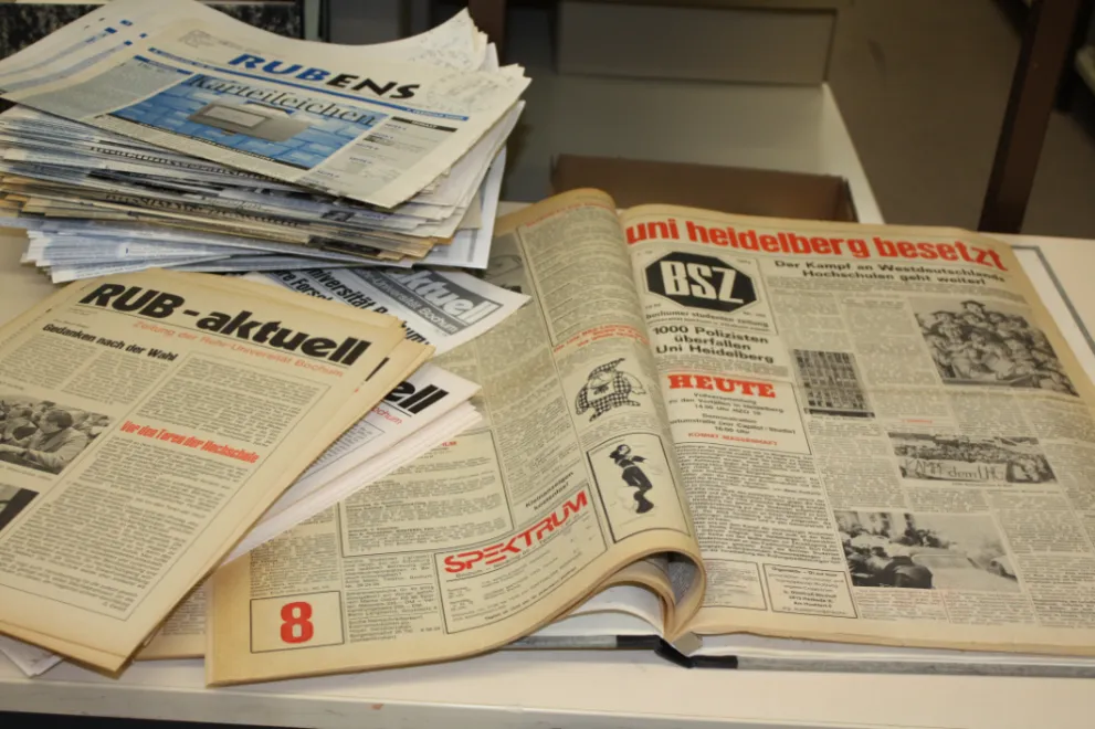 Auf einem Tisch liegen auf der linken Seite zwei Stapel Zeitschriften, RUBENS und RUB-aktuell, rechts daneben liegt ein aufgeschlagenes Exemplar der BSZ - Bochumer Studierendenzeitung