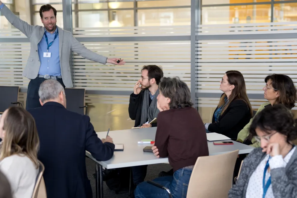 Workshop: Mehrere Personen sitzen an Gruppentischen und hören einem Vortagenden zu.