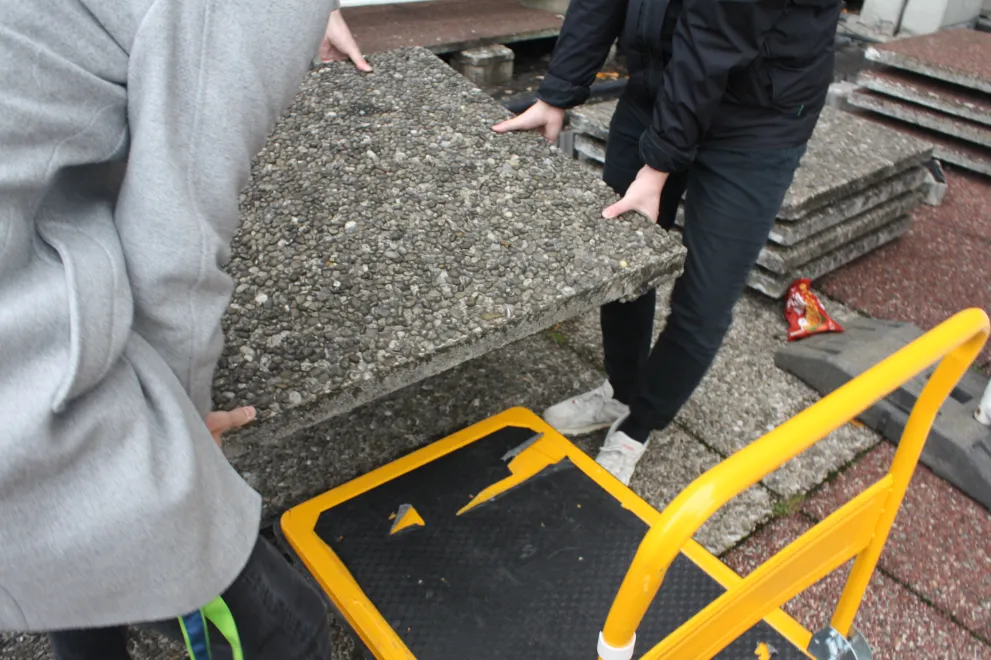 Zwei stark abgeschnittene Personen heben eine Waschbetonplatte, wie sie für die RUB typisch sind, an. Unten rechts im Bild steht bereits ein gelber Handkarren bereit um die Platte abzutransportieren.