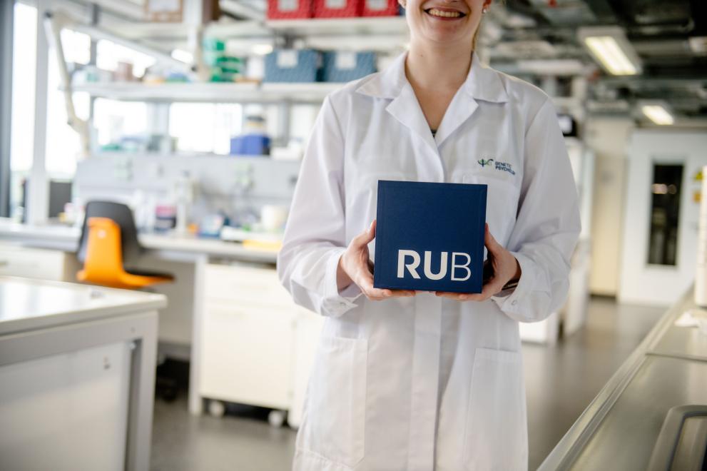 Foto: Frau im weißen Kittel steht in einem Labor und hält lächelnd einen RUB-Kubus vor sich. 