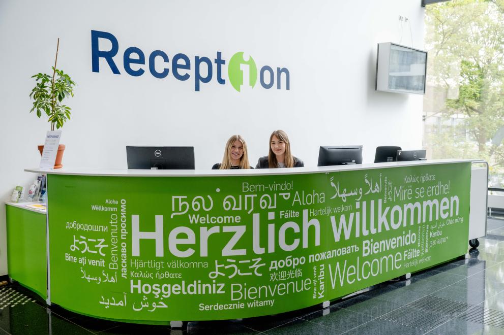 Photo: Two seated people and three monitors can be seen behind a green counter with the white sign "Welcome" in many different languages and fonts. There is a plant on the left end of the counter. On the wall is the lettering "Reception" and there is a monitor hanging on the wall.