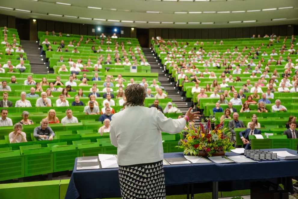 Photo: Well-filled lecture hall with green benches. The Chancellor stands at the front, a large bouquet of flowers next to her.