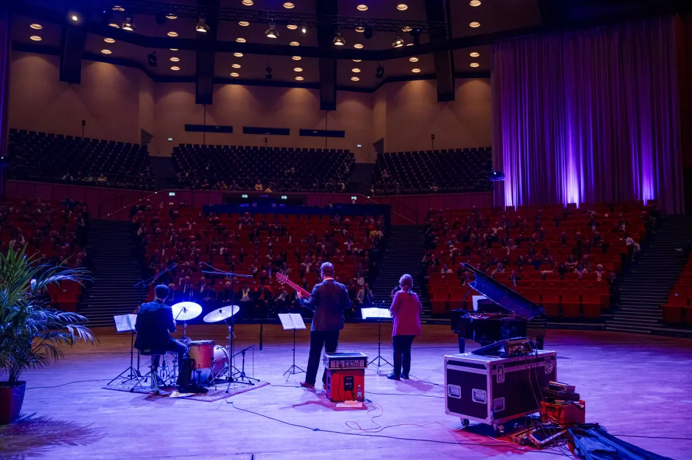 Foto: Band vor den Sitzreihen im Audimax. Der Saal ist abgedunkelt.