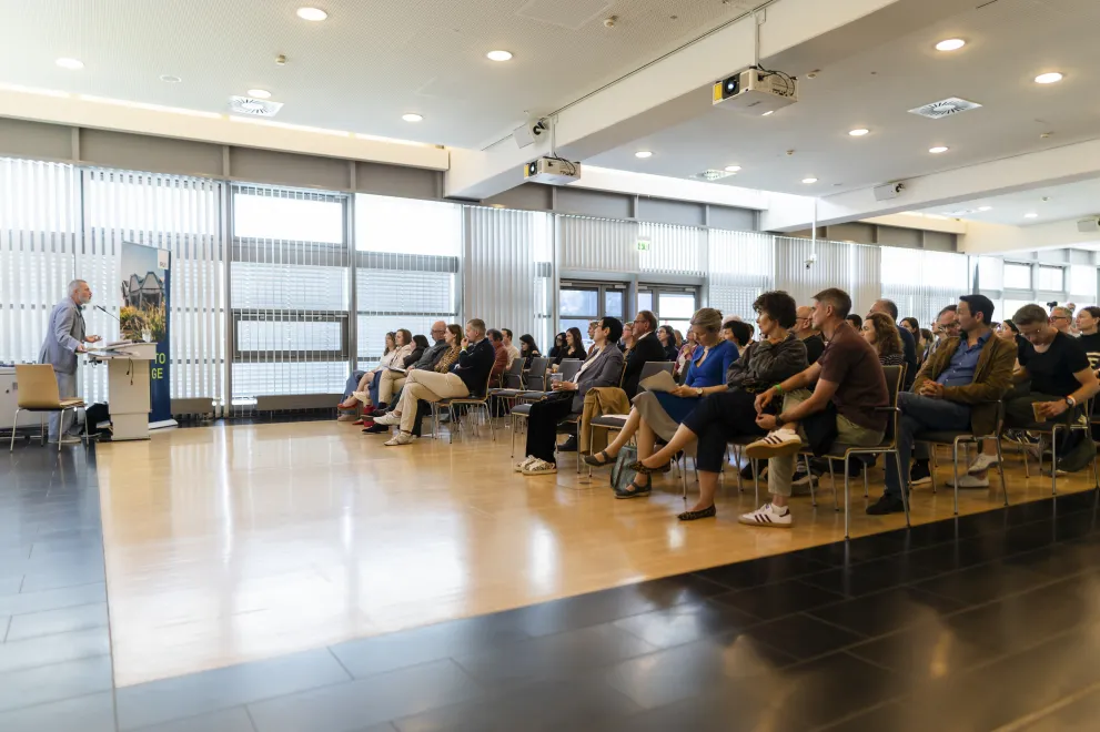 Foto: Vortrag im Veranstaltungszentrum. Eine Person steht hinter dem Rednerpult, die anderen sitzen und gucken die Person an.