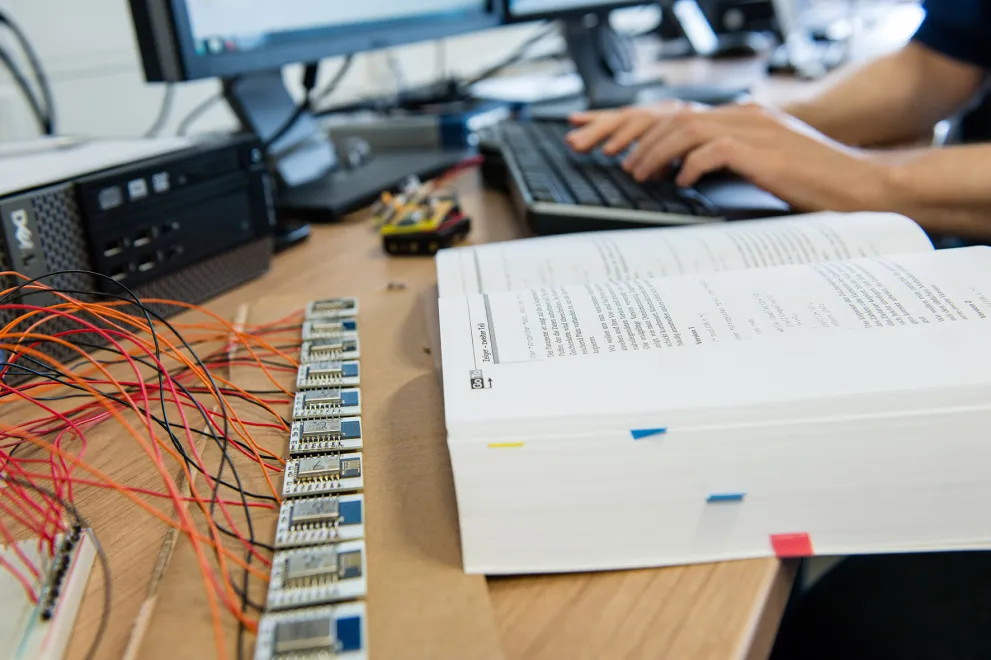 Foto: Auf einem Schreibtisch befindet sich ein aufgeschlagenes Buch, Platinen und Kabelgewirr sowie im Hintergrund auf einer PC-Tastatur tippende Hände.pende 