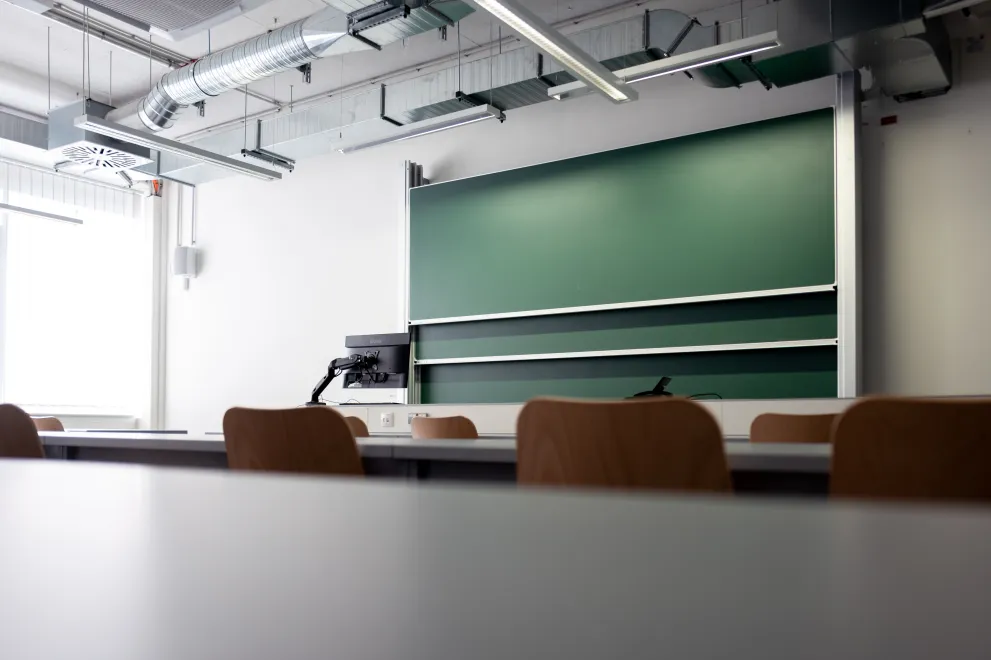 Foto: Seminarraum, vom Tisch aus fotografiert mit Blick nach vorne zu den Tafeln und einem Monitor.