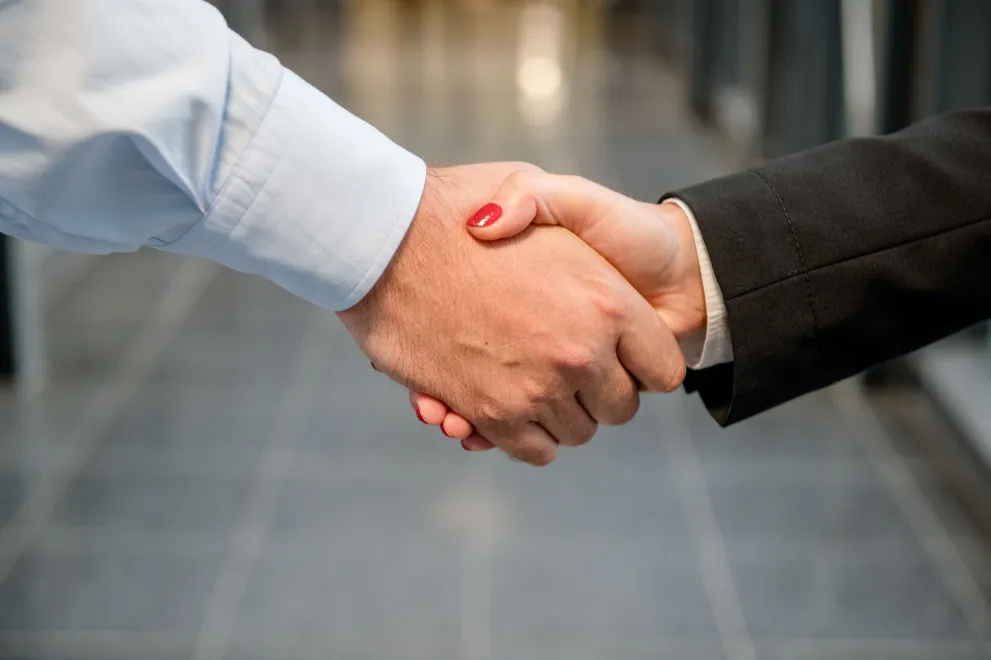 Das Bild zeigt einen Handschlag zwischen einer kräftigeren Hand mit Hemdsärmel und einer Hand mit einem schwarzen Blazer-Ärmel und rot lackierten Nägeln