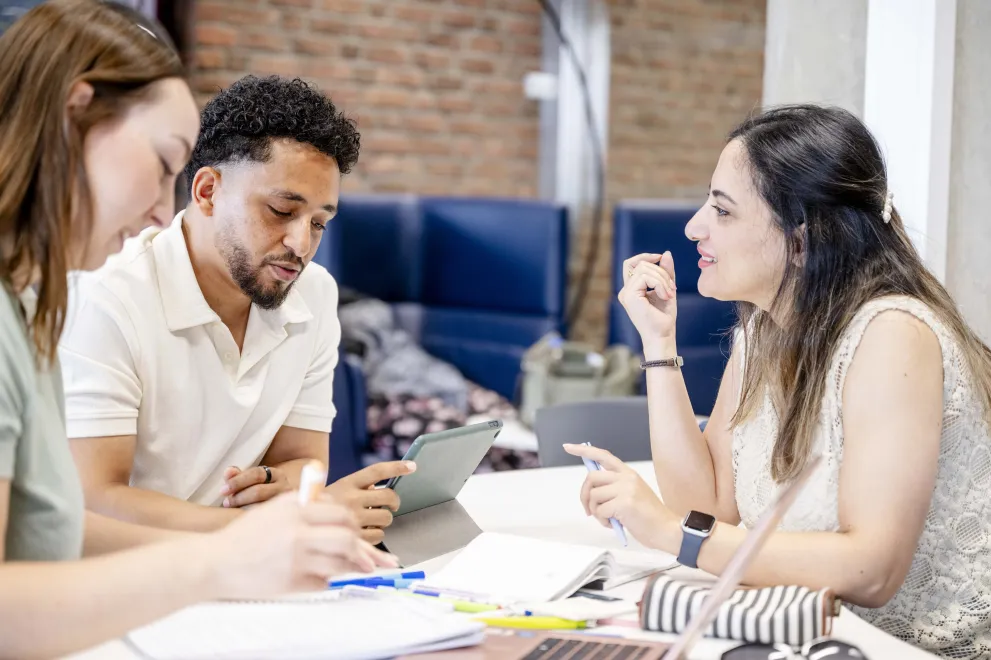 Drei Studierende sitzen an einem Tisch. Sie schauen auf ein Tablet und unterhalten sich. 