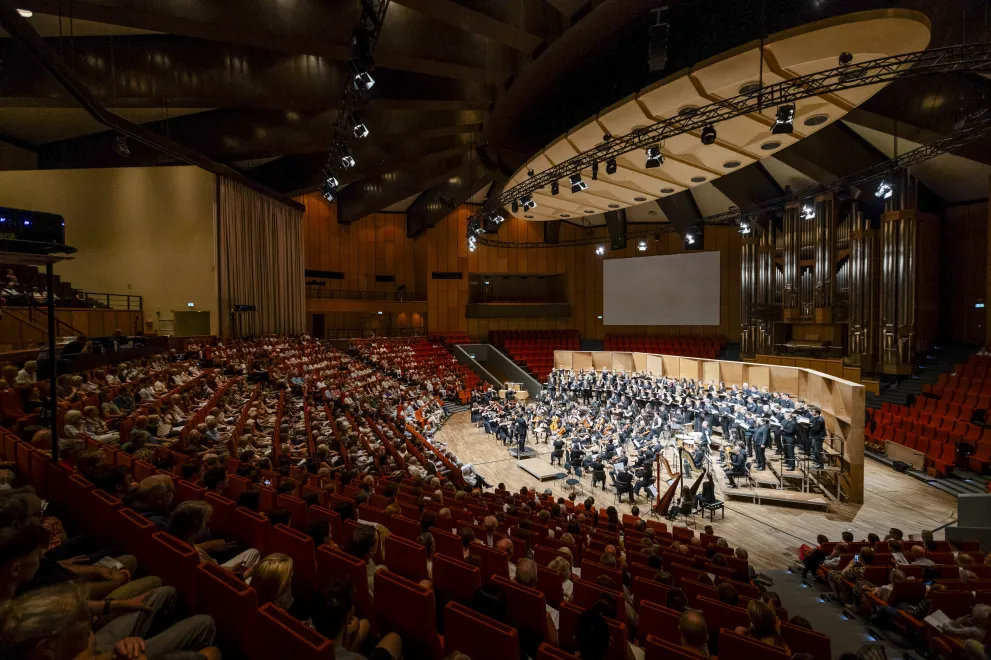 Foto: Blick aus den oberen Rängen des Audimax auf die Bühne mit dem Chor und Orchester des Semesterkonzerts 2026. Die Bühne ist beleuchtet. Die gut besetzten Sitzränge sind abgedunkelt.