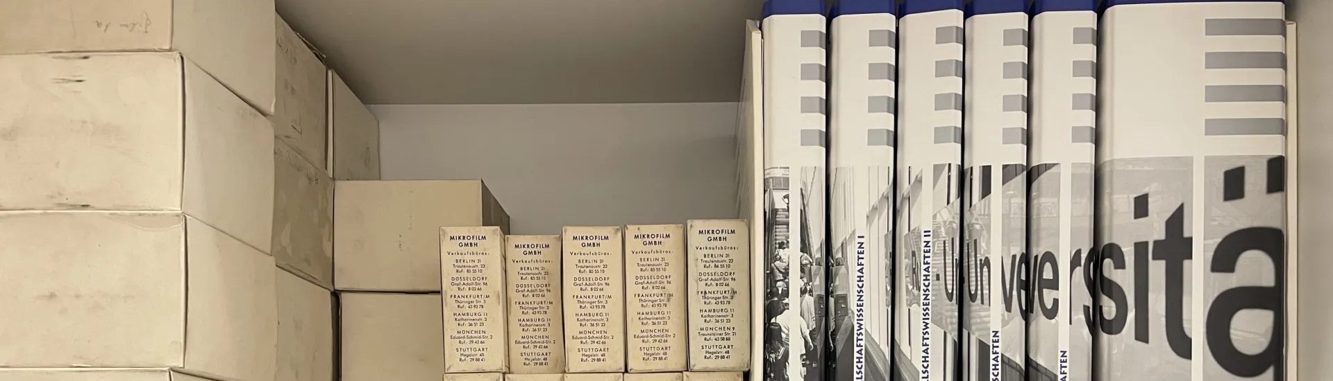 Auf zwei Regalbrettern befinden sich diverse Diasammlungen in transparenten Boxen, eine Publikationsreihe, deren Buchrücken zusammen ein Foto, auf dem der Schriftzug "Universität" zu sehen ist, ergeben, sowie diverse kleinere Materialien in Kartonagen zu sehen.