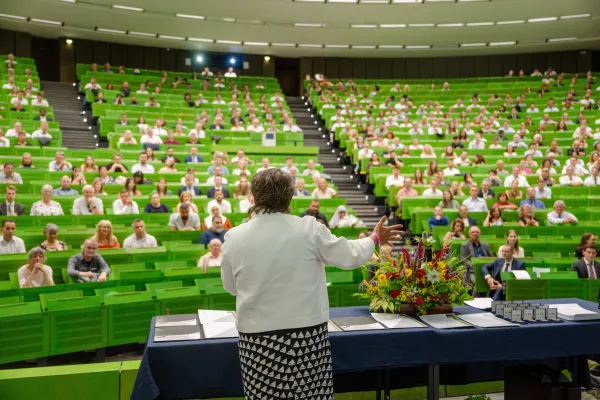 Photo: Well-filled lecture hall with green benches. The Chancellor stands at the front, a large bouquet of flowers next to her.