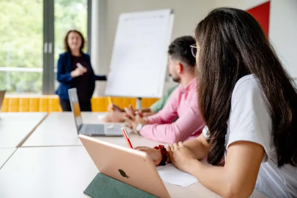 Foto: Seminar in einem Seminarraum. Zwei Personen sitzen und schauen zur Seminarleitung, die an einer Flipchart steht.